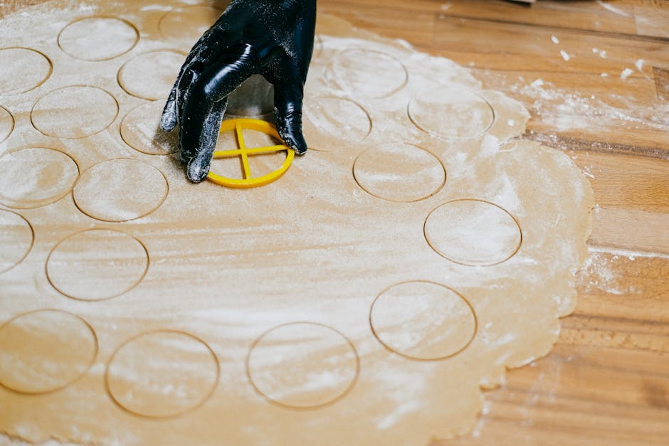 A gloved hand using a cookie cutter to shape dough on a floured surface.
