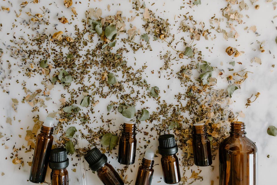 Glass bottles with essential oils surrounded by scattered dried herbs on a white surface.