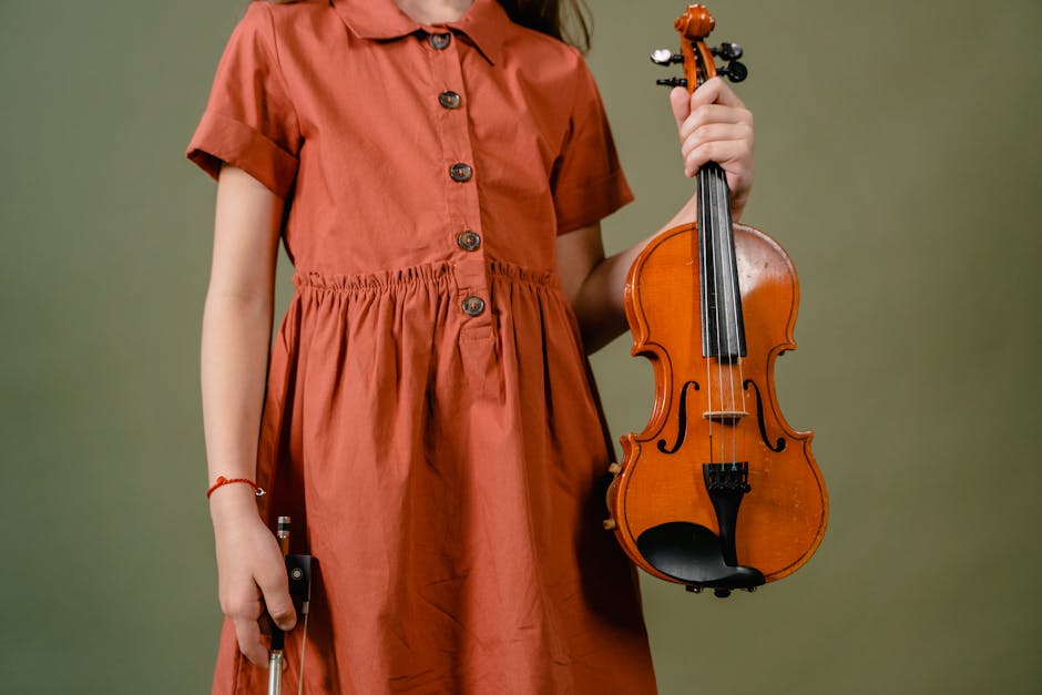 A young girl in an orange dress holding a violin against a green background, showcasing musical talent.